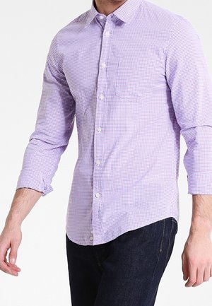 Hombre con una camisa de manga larga de cuadros morados y blancos abotonada, y jeans azul oscuro, de pie contra un fondo liso.