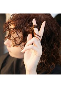 Lockigt, medellångt, rödbrunt hår med en glänsande textur. En hand med ljus hud håller i en liten hårsektion, fastsatt med en ring.