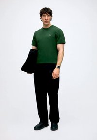 Joven con camiseta verde, pantalones negros, zapatos negros, sujetando una chaqueta negra, de pie frente a un fondo blanco liso.