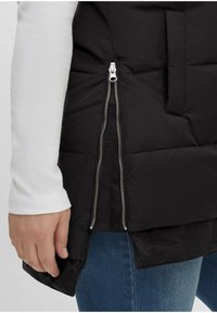 Schwarze Daunenweste mit seitlichem Reißverschluss und einer Stofftasche; getragen über einem weißen Langarmoberteil. Der Stoff erscheint glatt und leicht glänzend.