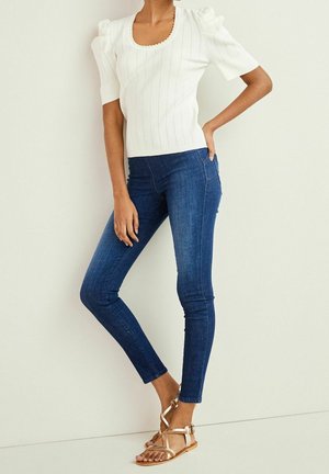 Femme portant un haut en maille blanc à manches courtes bouffantes, un jean skinny bleu foncé et des sandales plates dorées métalliques, debout contre un mur clair.