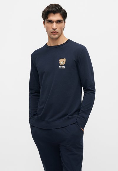 Άνδρας με σκούρα μαλλιά φοράει μακρυμάνικο πουκάμισο σε ναυτικό μπλε και παντελόνι, το πουκάμισο έχει μικρό λογότυπο αρκουδάκι με το κείμενο "Moschino Underbear".