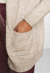 Hand i fickan på en beige stickad tröja, tillsammans med mörkröda läderbyxor som syns på sidan.