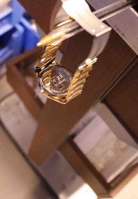 Gouden horloge met een metalen armband, met een ronde kast. Achterkant toont een gravure met tekst. Soepele textuur, gepolijste afwerking.