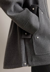 Manteau en mélange de laine gris avec une grande poche plaquée, des boutons-pression et des accents noirs contrastants sur la poche et la fermeture.