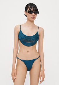 Teal bikini-set med en sammensydd bandeau topp med et gjennomsiktig panel og en lavt sittende bikiniunderdel. Glatt tekstur og minimale hardware detaljer.