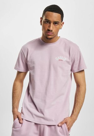 Junger Mann mit kurzen Haaren, der ein hellviolettes T-Shirt und passende Shorts trägt, steht mit den Händen in den Taschen vor einem einfarbigen Hintergrund.