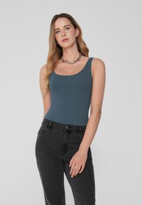Jonge vrouw met lang blond haar die een blauwe tanktop, zwarte jeans, zilveren ketting en ringetjes oorbellen draagt tegen een effen achtergrond.