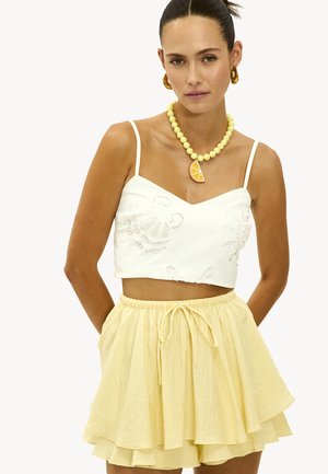 Mujer con top corto blanco bordado, falda de capas amarilla clara, collar de cuentas amarillas con colgante de rodaja de naranja y pendientes de aro dorados.