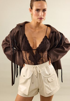 Femme portant un body en dentelle marron foncé, un short crème à liens avec poches et une veste marron avec de longues franges.