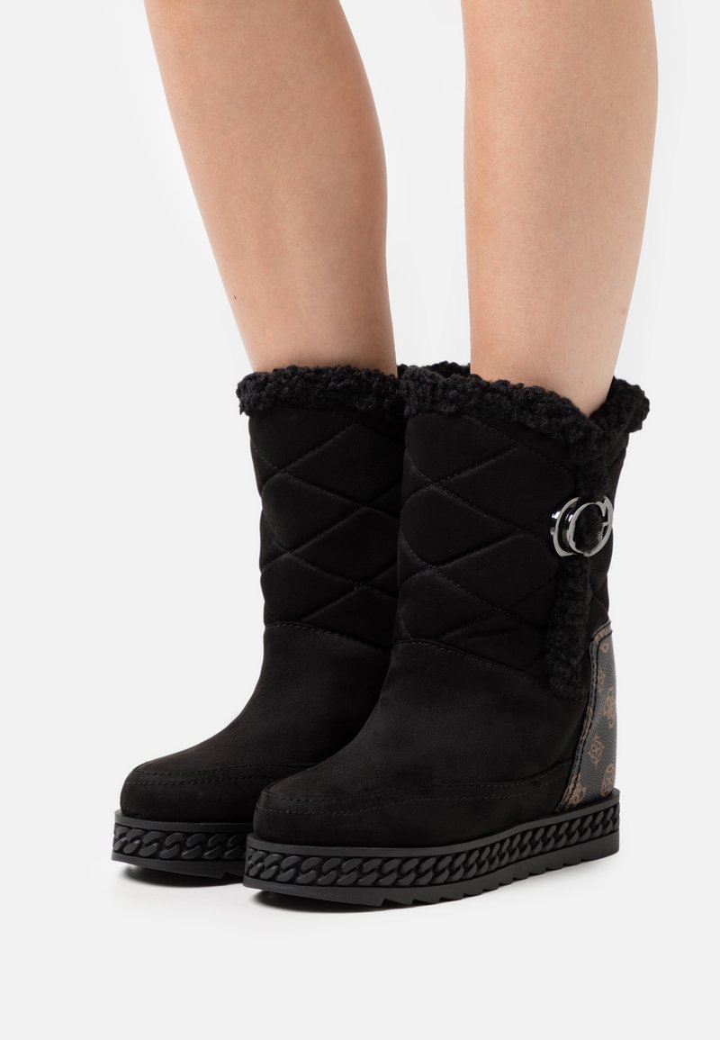 Bottes d’hiver noires mi-mollet en tissu matelassé, bordure en fausse fourrure, boucle latérale, panneau de talon à motifs et semelles épaisses texturées sur jambes nues.
