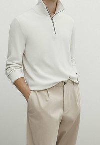 Pull côtelé crème clair avec col demi-zip, associé à un pantalon plissé beige. Le pull a des manches longues et un tissu texturé.