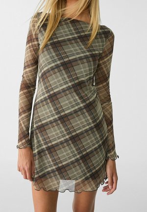 Femme portant une robe courte à manches longues à carreaux dans des tons de vert et de marron, avec un tissu légèrement transparent et un ourlet festonné.