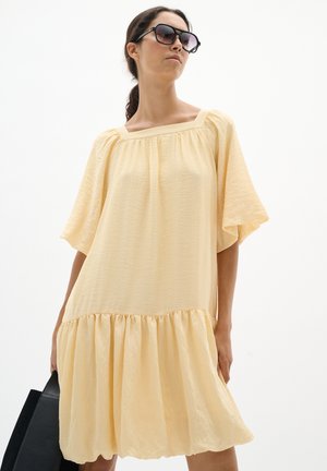 Kobieta ubrana w luźną jasnożółtą sukienkę z szerokimi rękawami i kwadratowym dekoltem, trzyma czarną torebkę i nosi duże czarne okulary przeciwsłoneczne.