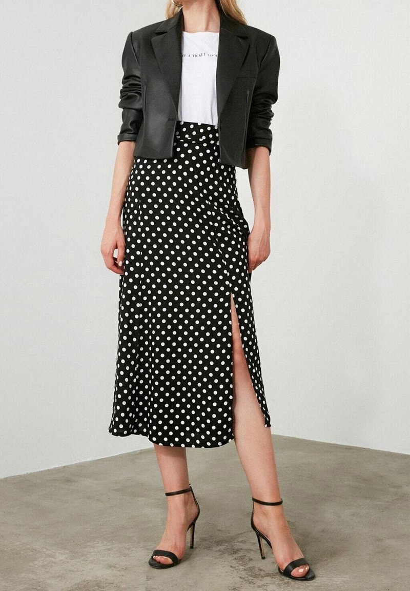 Femme portant un blazer noir court, un t-shirt blanc, une jupe midi noire à pois blancs avec une fente à l'avant, associée à des sandales à talons hauts noires.