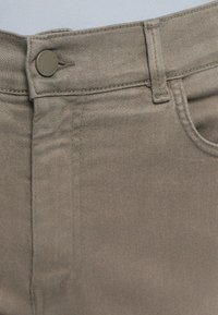 Nærbilde av beige denimbukser med midje, metallknapp og beltehempe på en lys bakgrunn.