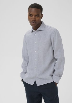 Mand med kort hår iført en lysegrå skjorte med knapper og lange ærmer samt mørke bukser, stående foran en ensfarvet lys baggrund.