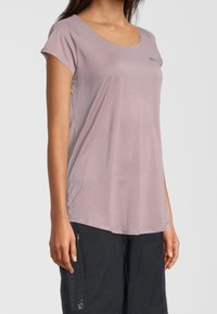 Kobieta w krótkim rękawie fioletowo-różowej koszulce sportowej i czarnych szortach z zamkiem błyskawicznym po boku, stojąca na jednolitym tle.