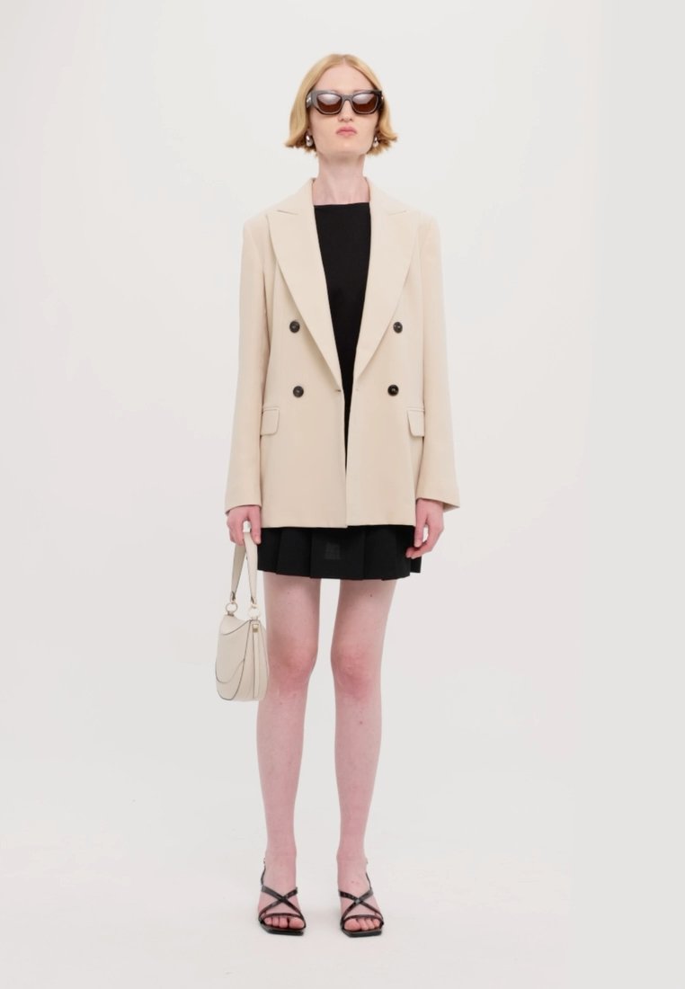 Blazer beige croisé avec des boutons noirs, associé à une robe noire, des sandales noires à lanières et un petit sac à main blanc.