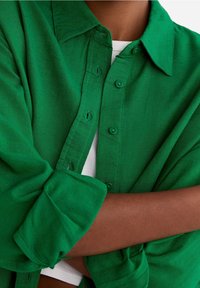 Chemise verte boutonnée en tissu texturé. Comprend un col, une patte de boutonnage avec des boutons et des manches retroussées. Portée par-dessus un vêtement blanc.