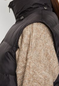 Svart nylon pufferväst med en glänsande yta, prydd med silverknappar, lagd över en ljusbrun stickad tröja med ett prickigt mönster.