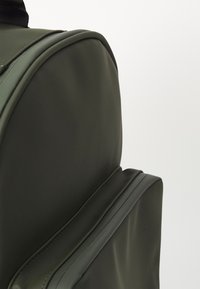 Sac à dos vert olive en matériau lisse, de forme arrondie, avec une poignée sur le dessus et une poche zippée sur le côté avec des coutures soignées.