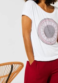 T-shirt bianca con un design circolare centrale caratterizzato da dettagli rosa e neri. Maniche corte, vestibilità rilassata, abbinata a pantaloni rossi texturizzati.