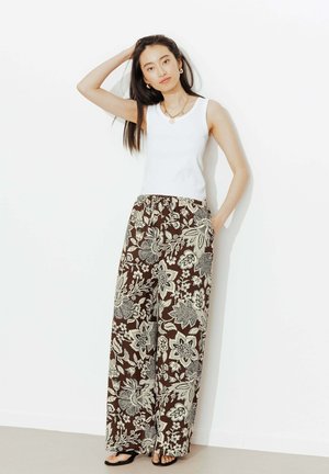 Femme portant un haut blanc sans manches et un pantalon large marron à motifs floraux, debout pieds nus contre un mur blanc une main dans la poche.