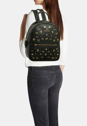 Donna che indossa uno zaino in pelle nero decorato con farfalle dorate, cuori, uccelli e motivi di mongolfiere, abbinato a una maglietta bianca e jeans scuri.