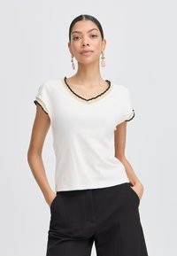 Top bianco a maniche corte con scollatura a V, caratterizzato da un bordo scallopato nero e oro attorno al colletto e alle maniche. Tessuto liscio, vestibilità casual.