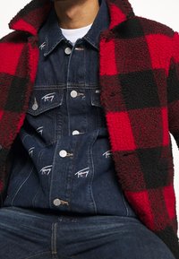 Röd- och svartrutigt fleecetak över en jeansskjorta med logotypdetaljer. Har knappstängning och en lager-på-lager-look med texturerade tyger.