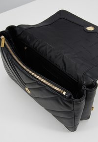 Musta nahkainen käsilaukku, jossa on tikkikuvio, kultaiset yksityiskohdat ja vetoketjukiinnitys. Sisäpuolella on kangaspinnoite, jossa on logokuvio.