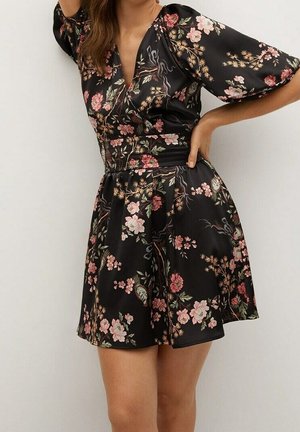 Femme portant une robe courte noire à motifs floraux roses et verts, manches bouffantes et décolleté en V, posant avec une main sur la hanche.