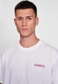 Fehér pamut póló, kerek nyakkal és rövid ujjakkal, bal mellen piros és kék "hummel" logóval.