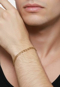 Gouden armband met schakels in ovale vormen, gedragen om een pols met lichte haren, tegen een neutrale achtergrond. Subtiele glans en textuur zichtbaar.