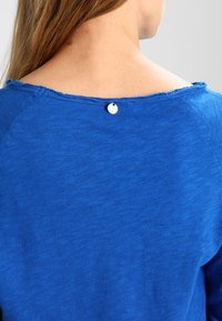 Maglietta a maniche lunghe blu con scollatura rotonda e bordi grezzi, decorata con un singolo bottone bianco sul retro, realizzata in un morbido tessuto testurizzato.