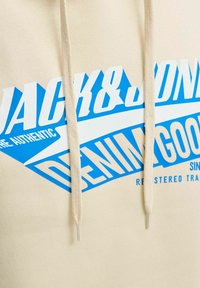 Krém színű kapucnis pulóver, nagy kék grafikával, amelyen a 'JACK&JONES' és a 'DENIM GOOD' felirat szerepel. Beige színű zsinórokkal, műanyag végződésekkel.