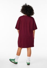 Bordo oversized majica haljina s kratkim rukavima, okruglim izrezom i glatkom teksturom. Uparena sa zeleno-bijelim tenisicama i bijelim čarapama.