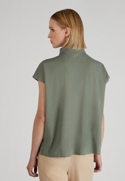 Kobieta z krótkimi blond włosami, ubrana w luźną, krótką oliwkową bluzkę i beżowe spodnie, widoczna z tyłu.