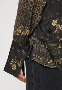 Camicia nera a maniche lunghe con motivi floreali e geometrici marroni, polsini abbottonati, indossata sopra jeans di denim scuri, mano appoggiata di lato.