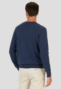 Hombre vistiendo un suéter de manga larga azul marino y pantalones beige, de pie con su espalda hacia el espectador contra un fondo claro y liso.