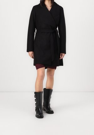 Zwarte wikkeljas met lange mouwen en een ceintuur, gecombineerd met zwarte leren laarzen tot aan de knie met gespen. Soepele textuur, minimalistisch ontwerp.