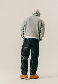 Persona vistiendo un suéter de punto con capucha gris, pantalones cargo de cuero negros y botas de trabajo color marrón, de pie con la espalda facing adelante.