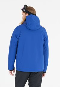 Giacca isolante blu con cappuccio, caratterizzata da una superficie liscia, polsini elasticizzati e una patch con logo sulla manica. Occhiali da sci neri sulla testa.