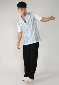 Chemise à manches courtes présentant un motif bleu avec des accents verts ; associée à un pantalon noir et des baskets blanches.