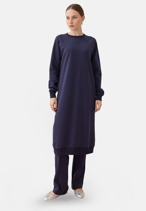 Femme debout portant une longue robe bleu marine par-dessus un pantalon assorti et des ballerines argentées, regardant légèrement vers sa gauche sur un fond blanc uni.