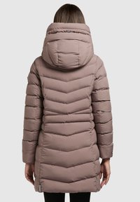 Femme de dos portant un long manteau d'hiver matelassé taupe avec une grande capuche et un motif de quilting en chevrons.