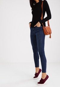 Svart ribbad långärmad topp, blå tajta jeans och burgundy mockaskor med en rosettdetalj. Brun axelväska med flätad rem.