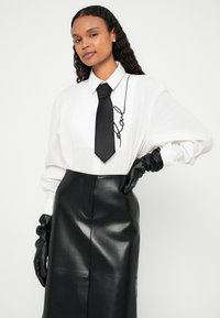 Camicia bianca con bottoni e maniche oversize, caratterizzata da una cravatta nera e un dettaglio iconico. Abbinata a una gonna nera in pelle a vita alta e guanti.