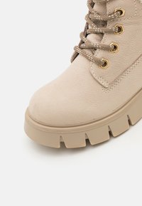 Beige kunstlær ankelstøvlett med avrundet tå, polstret krage og flat brune såle med dype spor. Guldfarget utstyr på snøringene.